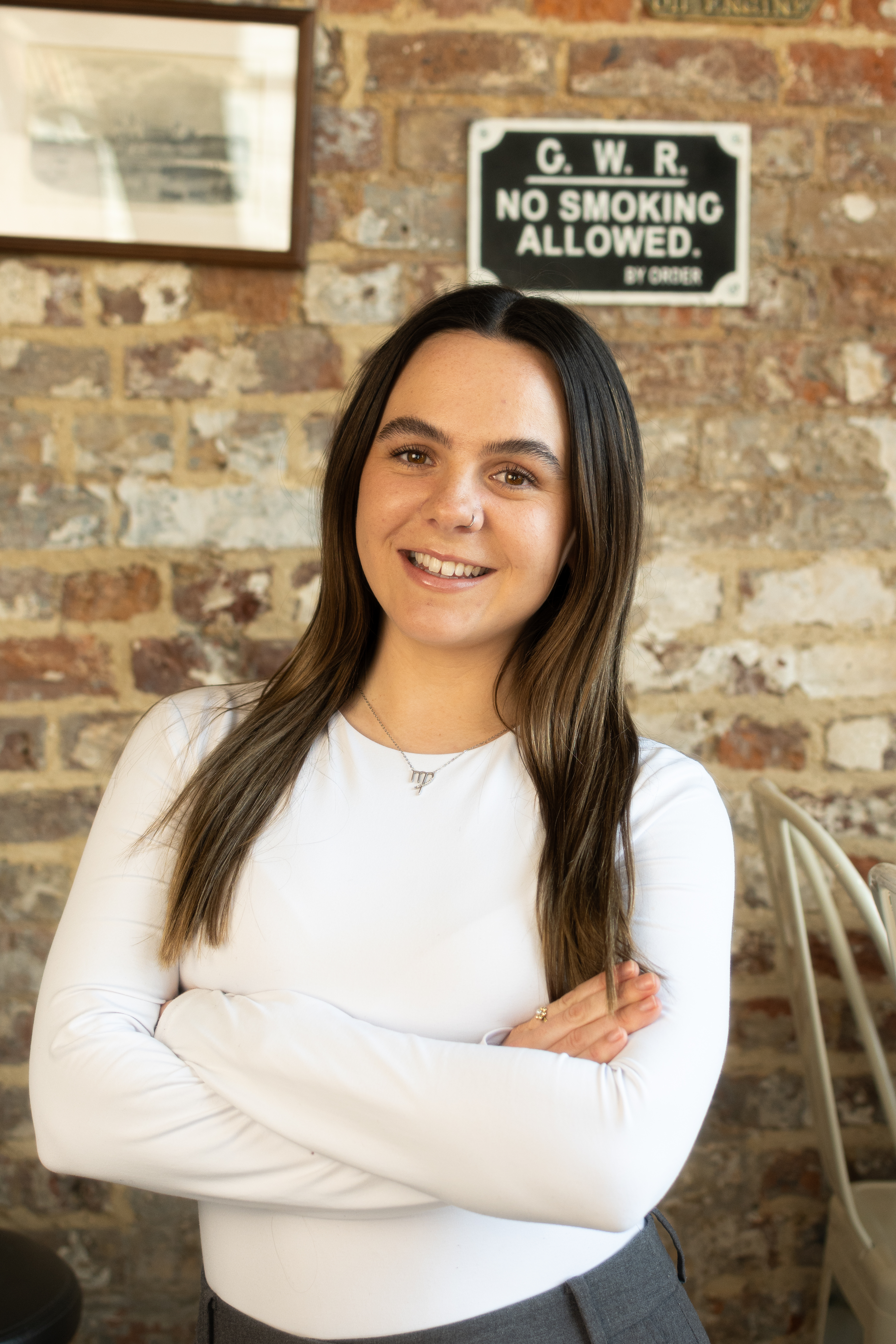 A woman with long dark hair, wearing a white shirt and standing in front of a wall with a sign and a framed picture.